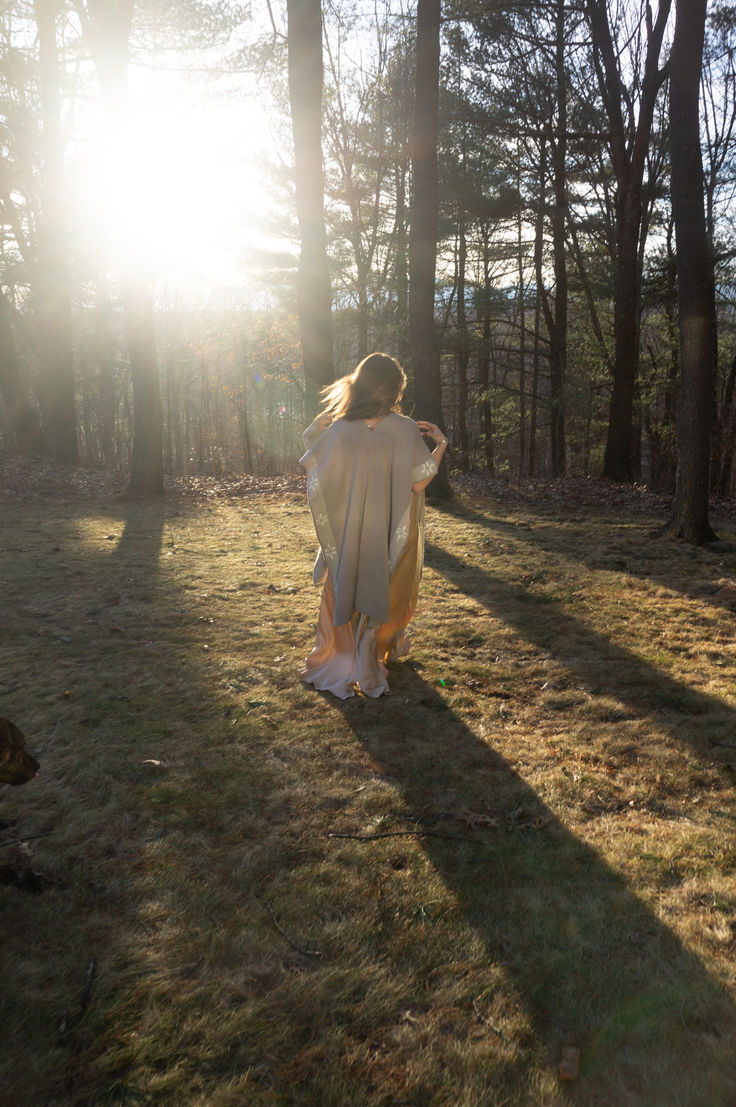 a woman is walking through the woods with her back to the camera and sun shining behind her