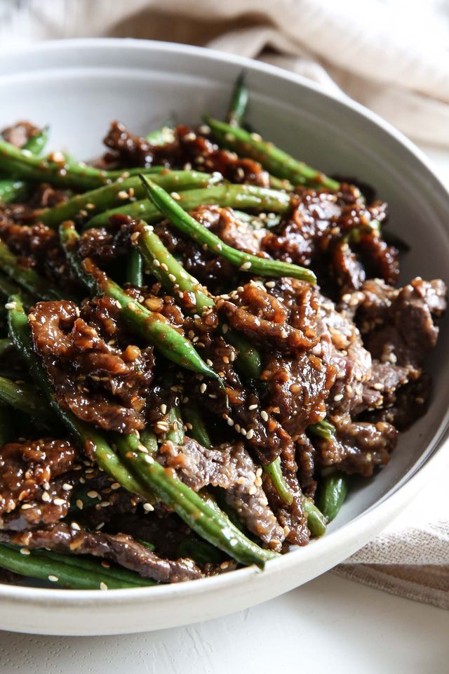 a white bowl filled with meat and green beans on top of a tablecloth next to a fork