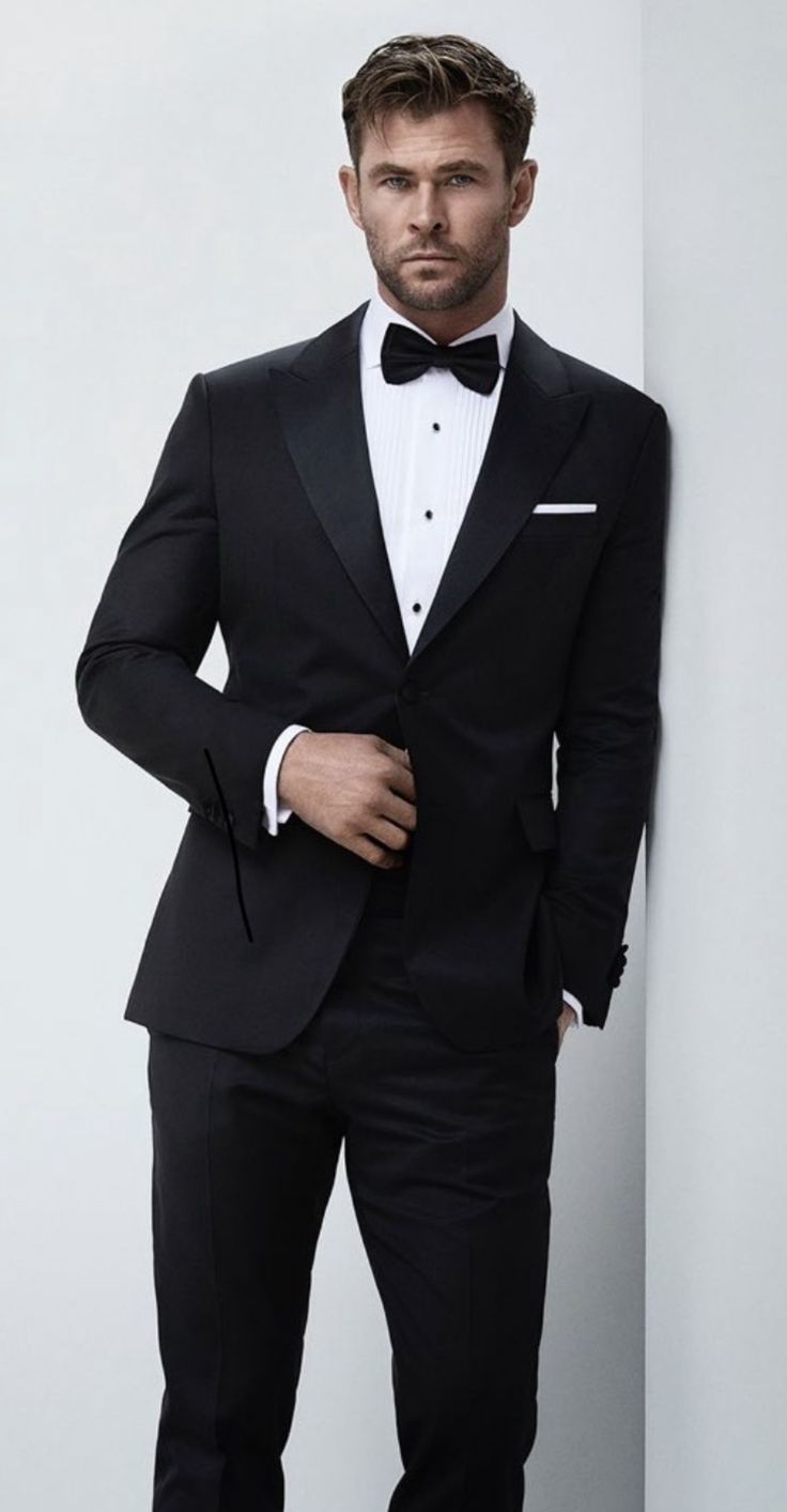 a man in a tuxedo leaning against a wall with his hands on his hips