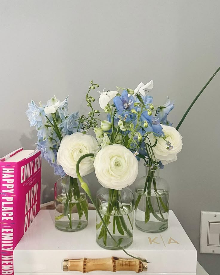 some white and blue flowers are in vases on a table next to a pink book