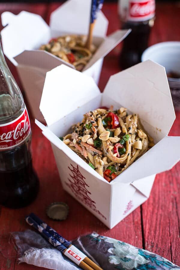 two take out boxes filled with noodles and veggies next to a bottle of coke