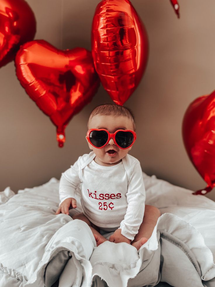 a baby wearing sunglasses and sitting on a bed with red heart balloons in the background