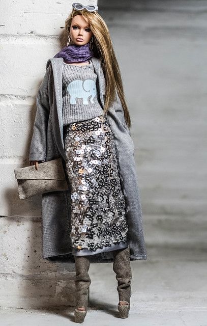 a barbie doll leaning against a white brick wall wearing a gray coat and sequin skirt