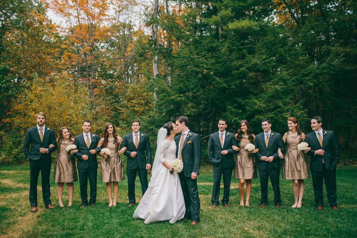 a bride and groom kissing in front of their wedding party outside on the grass with trees behind them