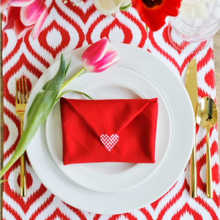 a place setting with red napkins and white plates, gold cutlery and pink tulips