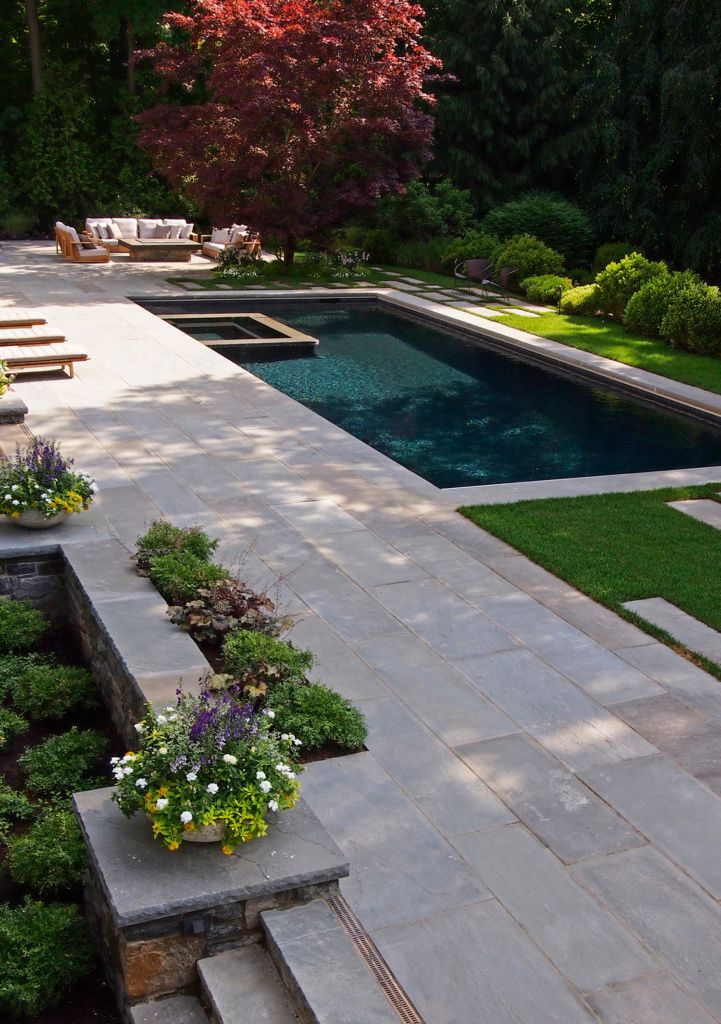 an outdoor pool surrounded by greenery next to a stone patio with benches and tables