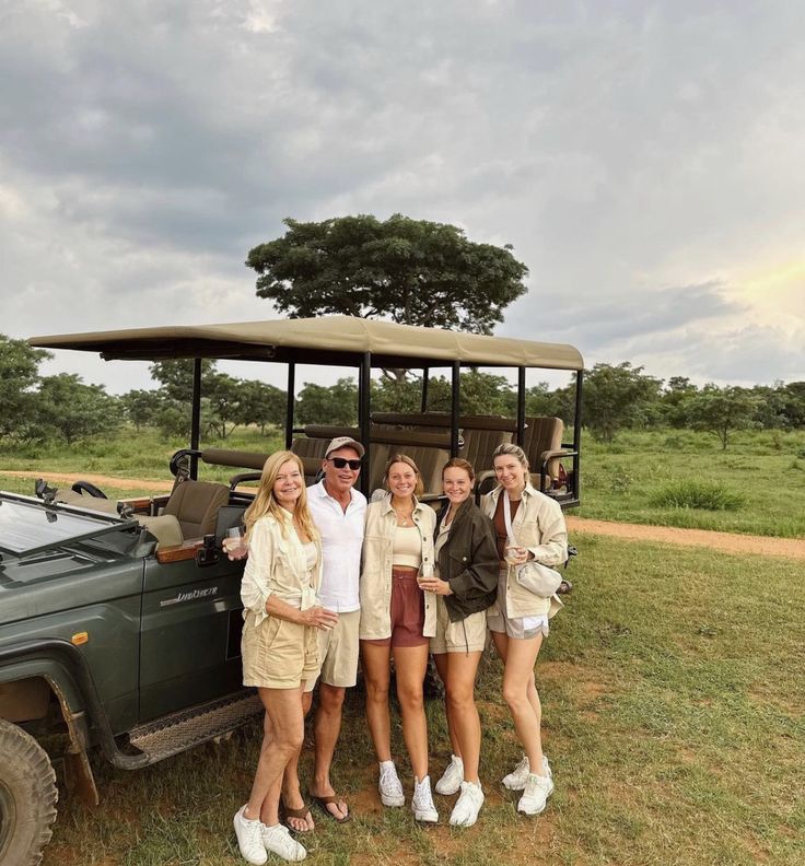 four women standing in front of a safari vehicle on the grass with an elephant behind them