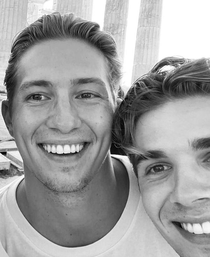 two men are smiling for the camera in front of the parthenion, one is wearing a white t - shirt