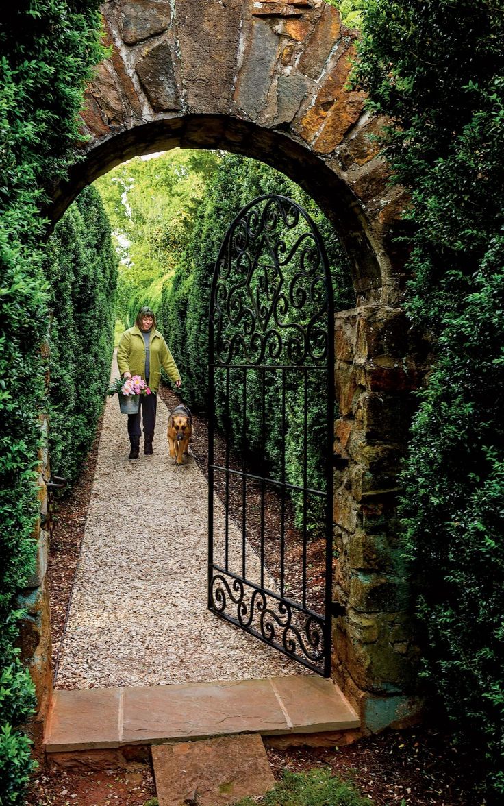 a man walking his dog through an iron gated entrance to a lush green garden