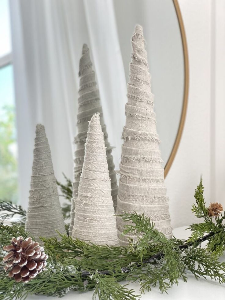 three white christmas trees sitting on top of a table next to a mirror and pine cones