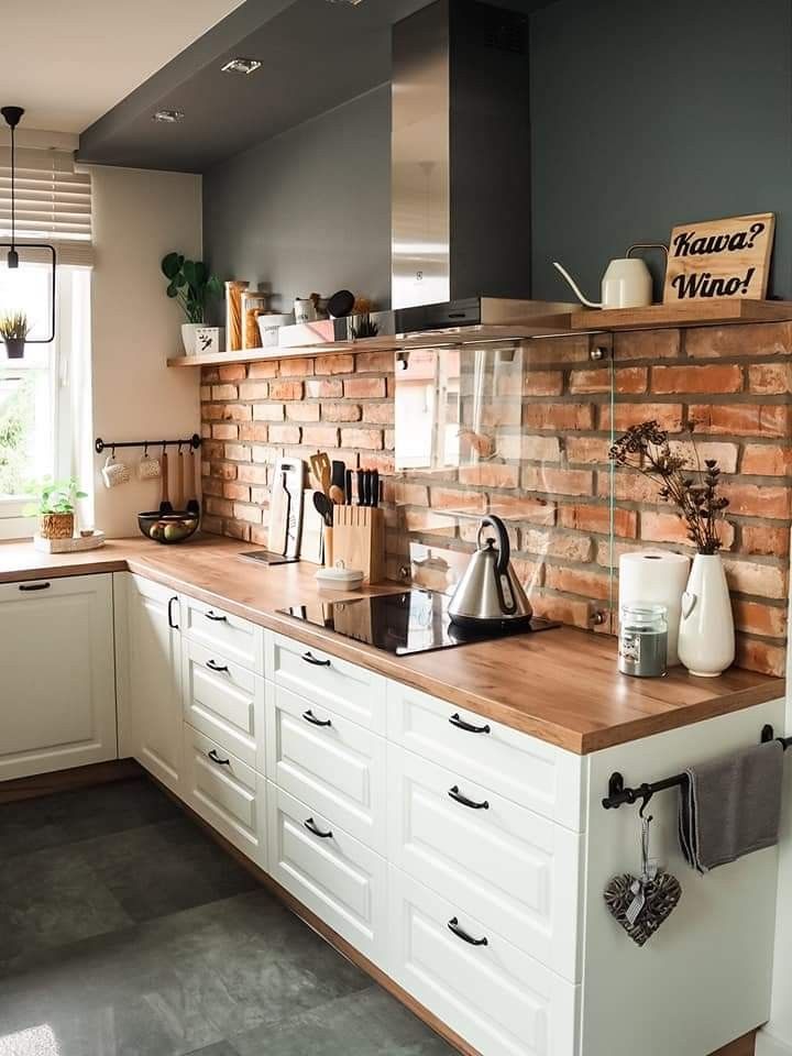 a brick wall in a kitchen with white cabinets and counter tops, along with a wooden cutting board