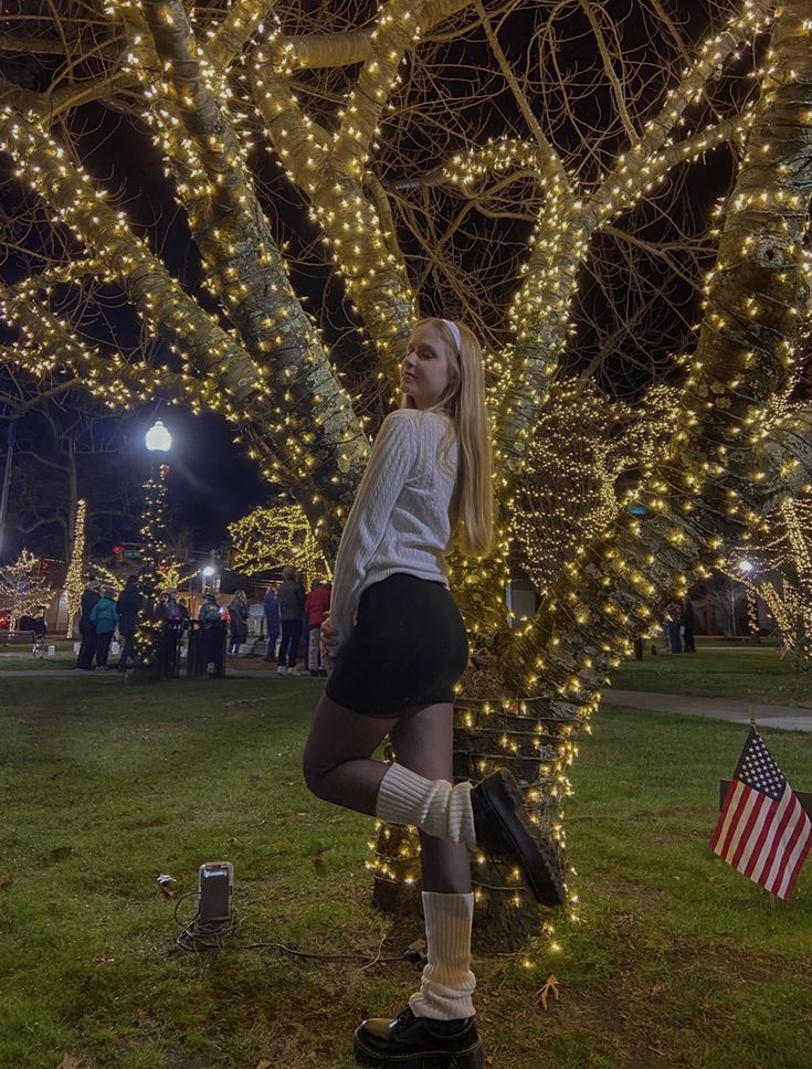 a woman standing in front of a tree with christmas lights all around her and an american flag on the ground