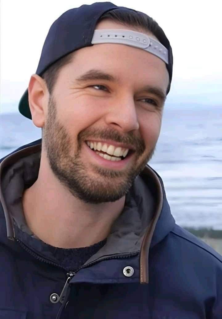 a man with a beard wearing a hat and smiling at the camera while standing next to the ocean