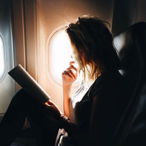 a woman sitting on an airplane reading a book and looking out the window at the sky