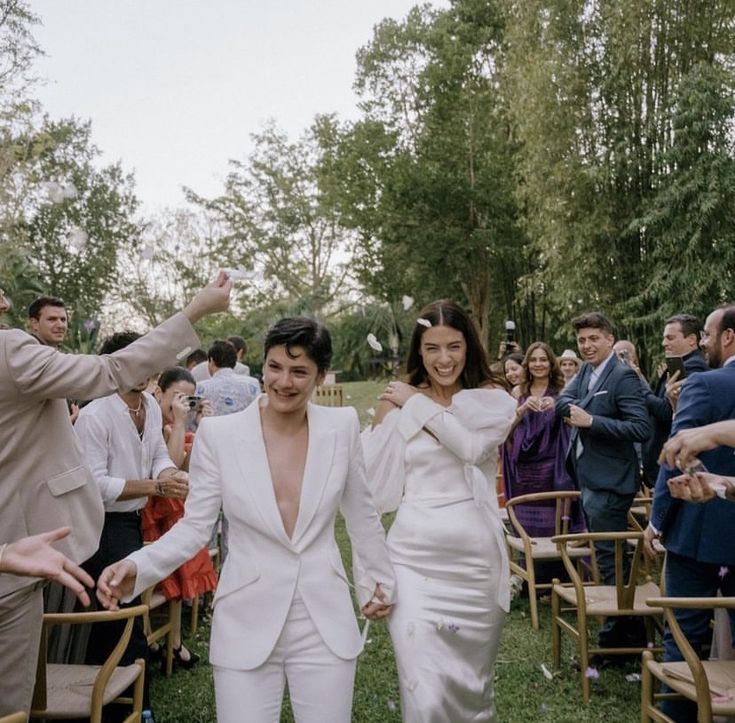 two people are walking down the aisle with their arms in the air and one person is wearing a white suit