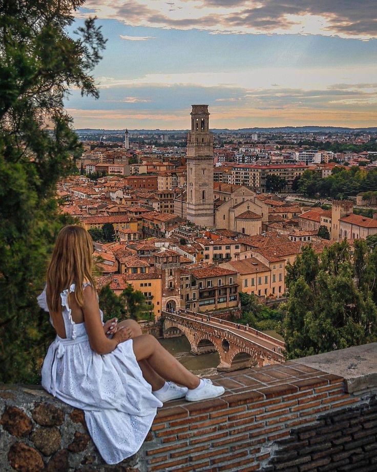 a woman sitting on top of a brick wall next to a tree and looking at the city