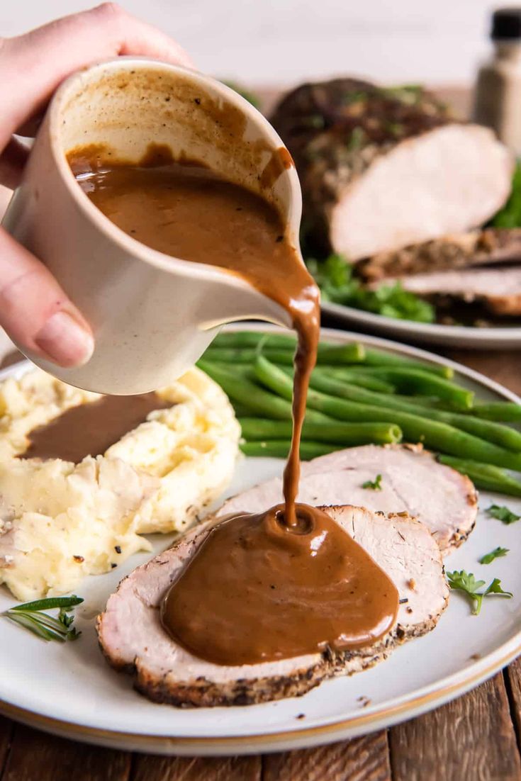 someone pouring gravy on some meat with mashed potatoes and green beans