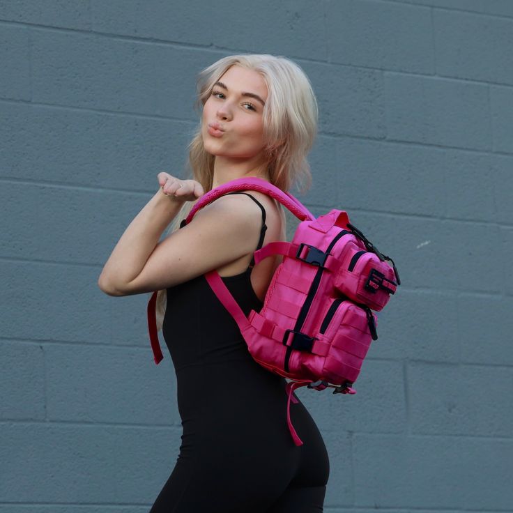 a woman with blonde hair wearing a pink backpack and black leggings stands in front of a gray brick wall