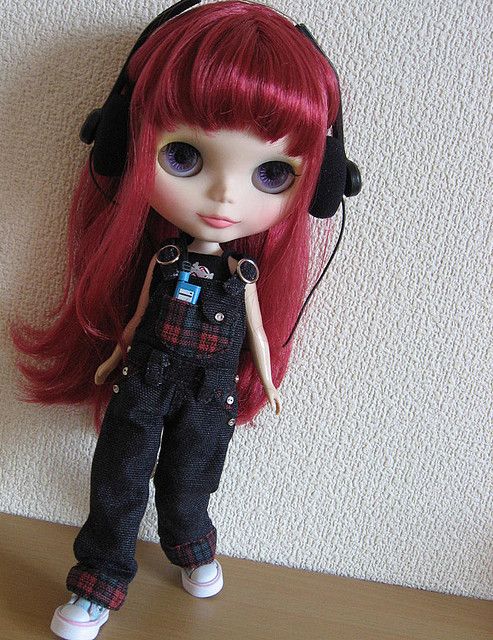 a doll with red hair wearing headphones and black overalls is standing on a table