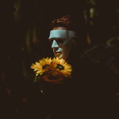 a man with sunflowers in his mouth and face painted white, standing next to trees