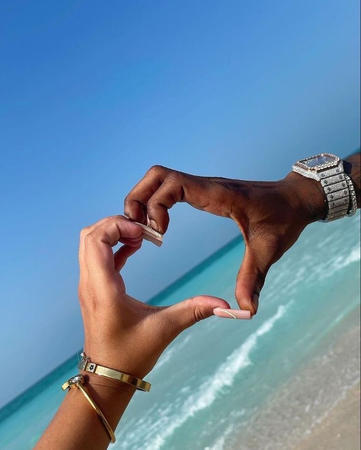 two hands making a heart shape with their fingers on the beach, one holding a cell phone