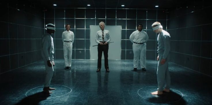 five men in white suits and blindfolds are standing in a dark room, looking at each other