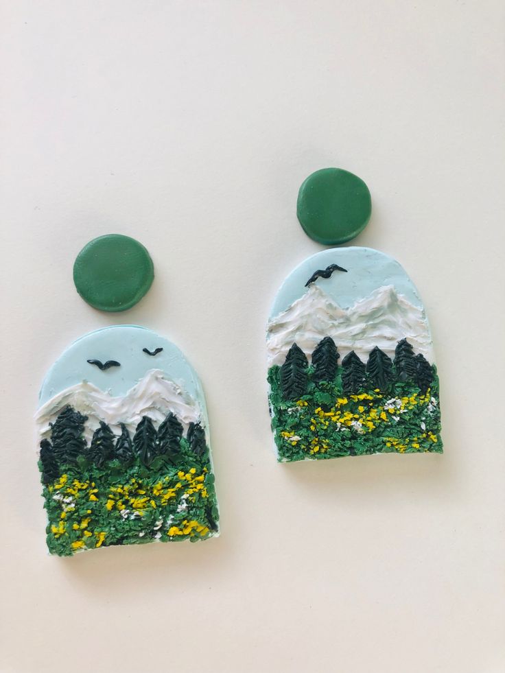 two green and white earrings with mountains in the background, one is made out of clay