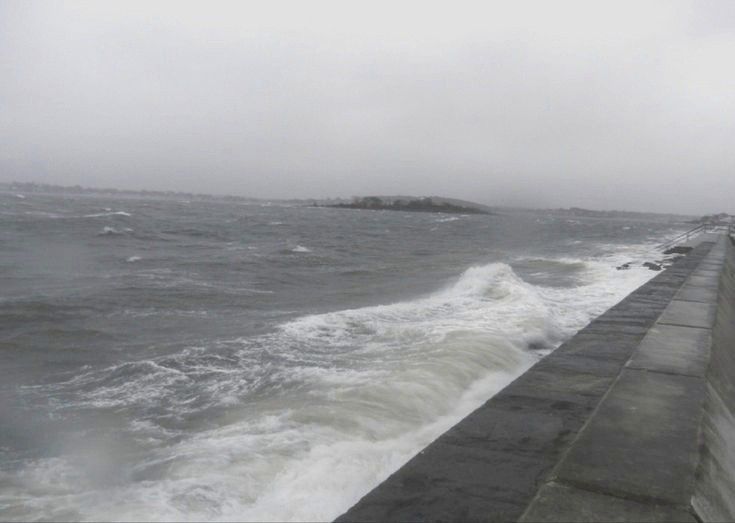 the ocean is choppy and stormy with waves crashing against the concrete wall, along with an island in the distance