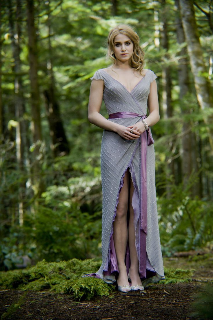 a woman standing in the middle of a forest wearing a dress with a purple sash