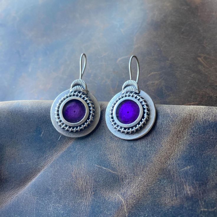 These Boho Silver Dangle Earrings feature a unique round tribal design with purple enamel at the center. The intricate pattern on each earring is complemented by a detailed ornate border, adding a touch of elegance and allure. The hooks ensure a secure and comfortable fit, making these earrings perfect for any occasion. Handcrafted from silver-plated Zamac, a material known for its durability and resistance to tarnish. The purple enamel is carefully selected for its vibrant color and smooth finish. Each earring measures 3.8 cm in length and 2.2 cm in width (approximately 1.5 inches by 0.87 inches). Every pair is meticulously handmade by me, ensuring a unique and personal touch in each piece. These earrings can be a perfect gift for your loved ones, offering both style and meaning. Care Ins Purple Fusion Style Round Jewelry, Nickel Free Purple Earrings For Festival, Traditional Purple Jewelry For Festival, Purple Traditional Festival Jewelry, Traditional Purple Drop Earrings, Nickel-free Purple Jewelry For Festivals, Bohemian Purple Round Hoop Earrings, Nickel-free Purple Earrings For Festival, Purple Round Metal Earrings