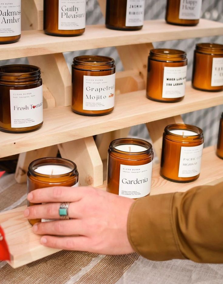 a person holding a candle in front of some jars on a wooden shelf with other candles