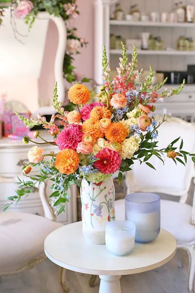 an arrangement of flowers in a vase on a table with two candles and some chairs