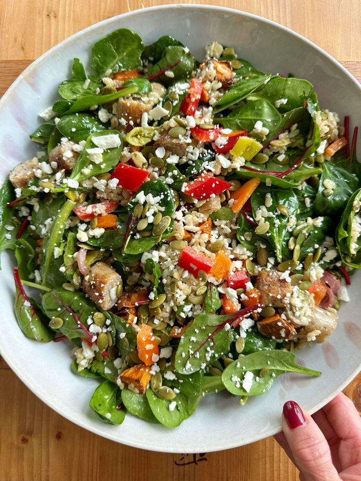 a white bowl filled with spinach salad on top of a wooden table next to a person's hand