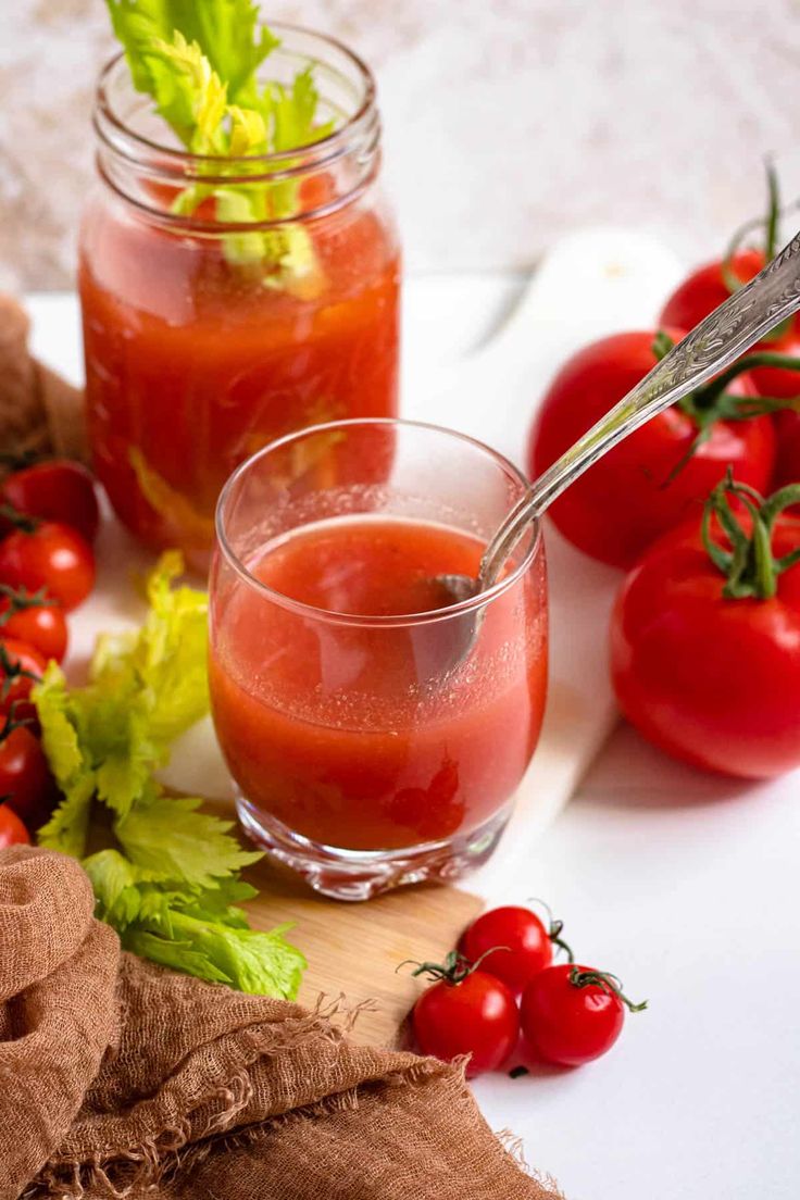 two glasses filled with tomato juice and garnished with fresh herbs, sitting on a cutting board