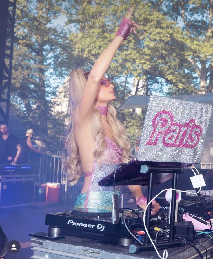 a woman with her arms up in the air at a music festival, while dj's are behind her