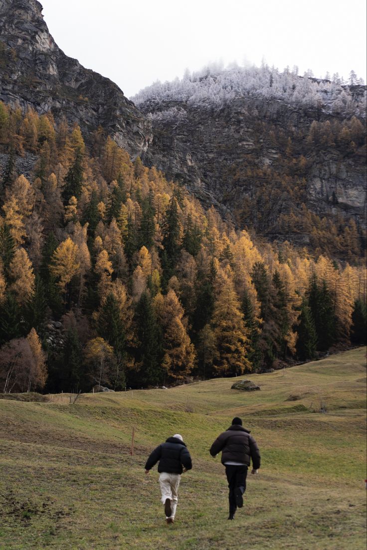 two people are running through the grass in front of some trees with yellow leaves on them