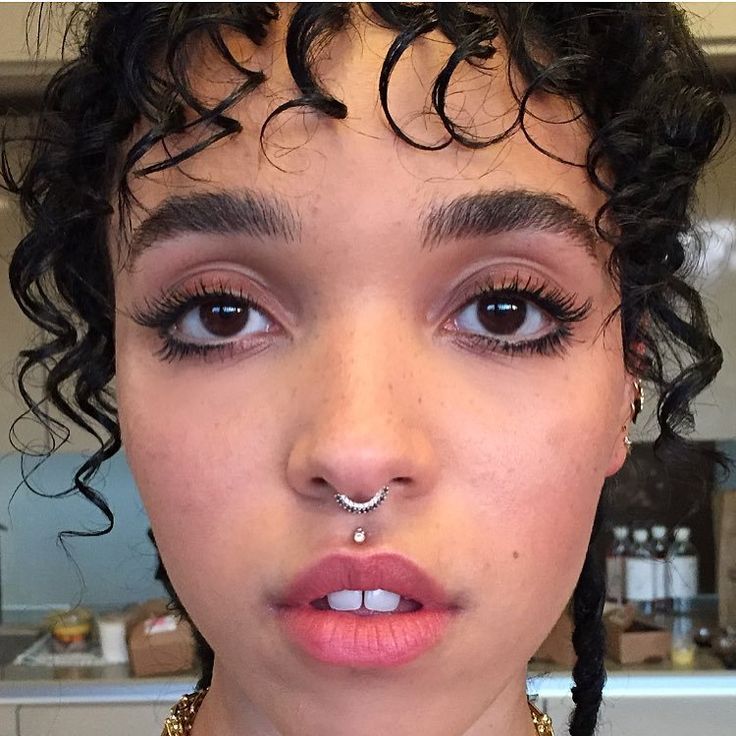 a close up of a person with curly hair and piercings on her nose wearing a necklace