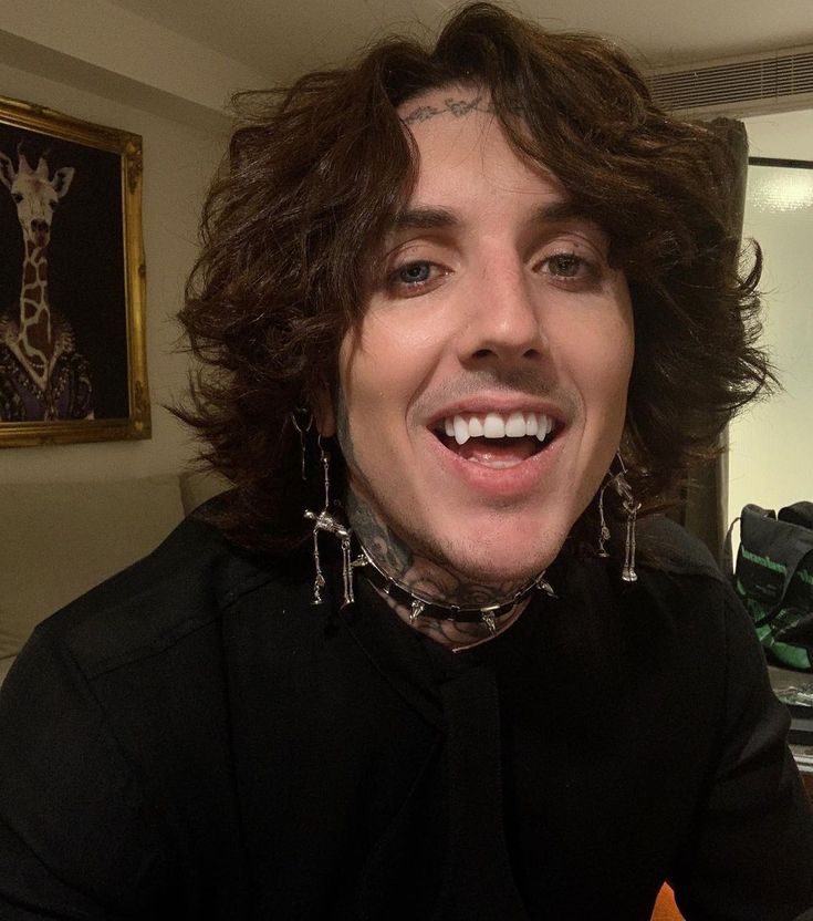 a man with long hair and piercings smiles at the camera while sitting in his living room