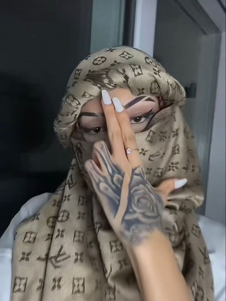 a woman with tattoos on her arm and hand covering her face, wearing a headscarf