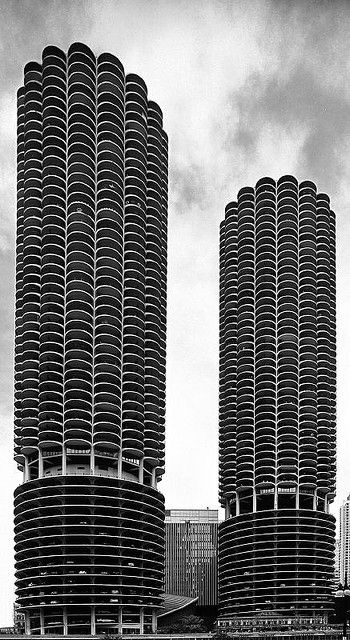 black and white photograph of two tall buildings in front of a body of water with boats on it