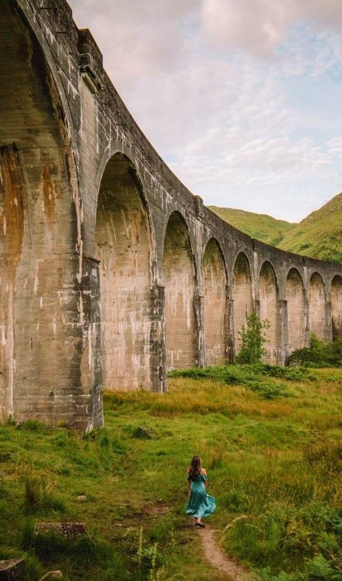 a woman standing in front of an old stone bridge with arches and grass on the ground