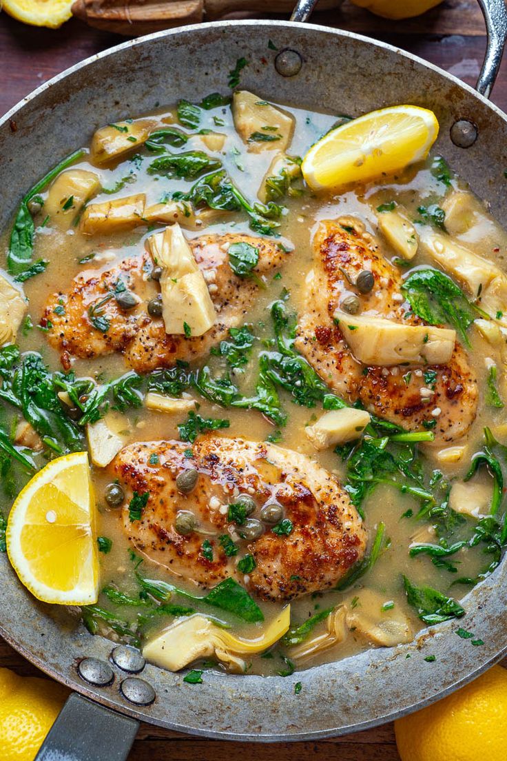 a skillet filled with chicken, spinach and lemons on top of a wooden table