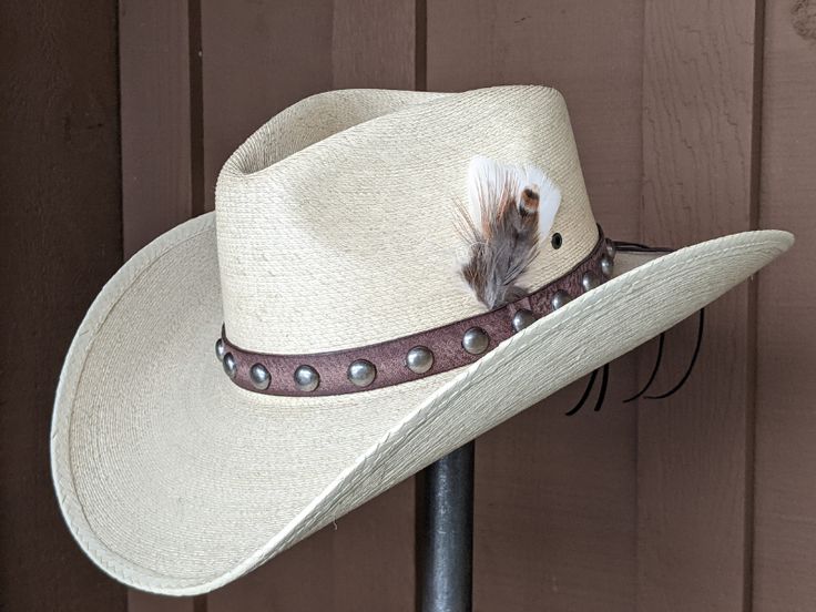 Chet starts with a Mexican soft palm leaf hat body that has a 3 3/4" brim and 5 1/4" crown. The band is simple with distressed brown leather and antique nickel color spots. The back of the band has a deer antler bead for accent. A neutral feather combo completes the hat. Note: Please allow 3-4 weeks. Rustic Adjustable Fedora With Flat Crown, Natural Color Adjustable Western Hat Bands, Adjustable Natural Color Western Hat Band, Adjustable Western Hat Band In Natural Color, Adjustable Straw Hat With Flat Crown For Country Events, Adjustable Natural Western Hat Bands, Fitted Rustic Hat For Country Events, Custom Adjustable Brown Hat Bands, Rustic Fitted Fedora For Country Events