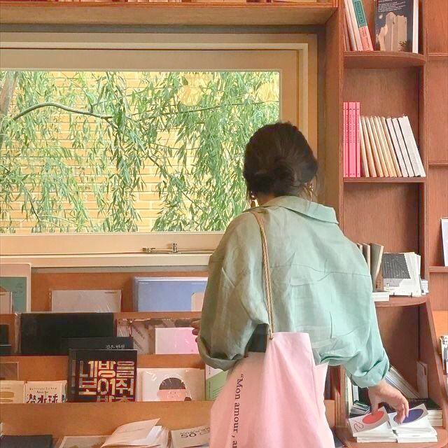 a woman standing in front of a bookshelf holding a bag and looking out the window