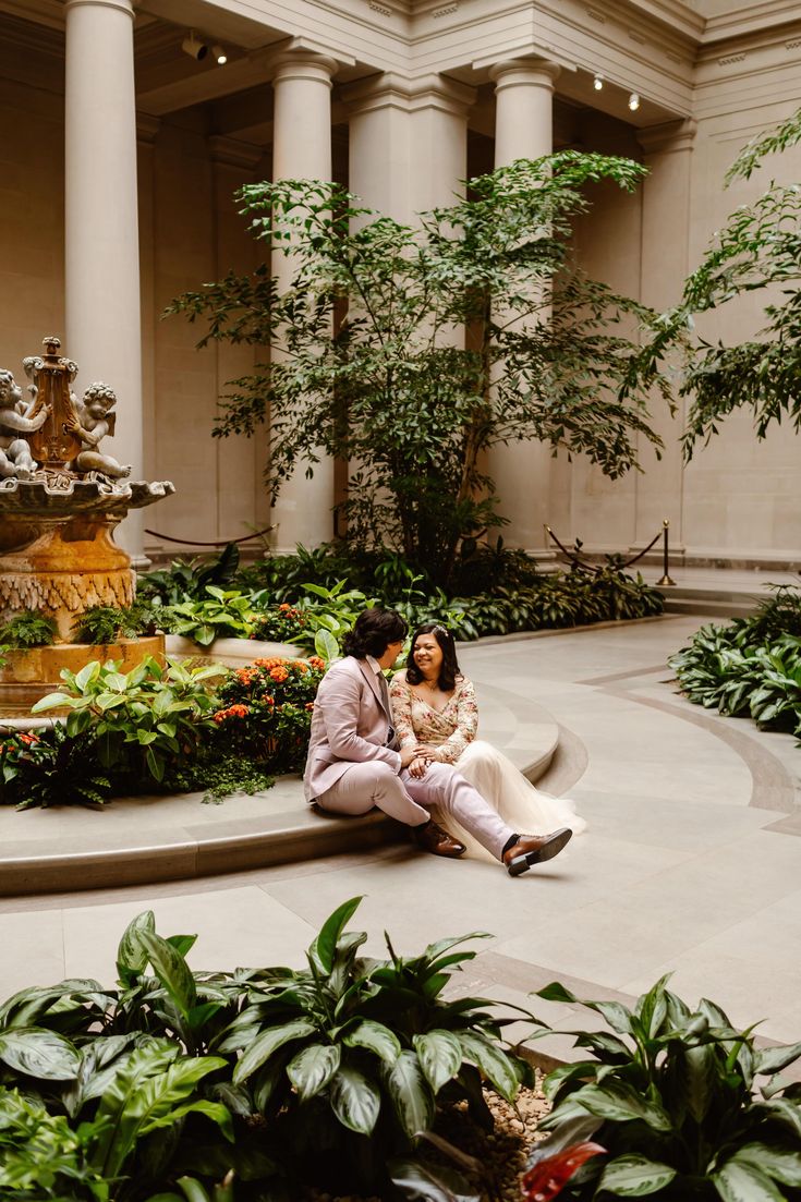 two people are sitting on the ground in front of some plants and water fountains