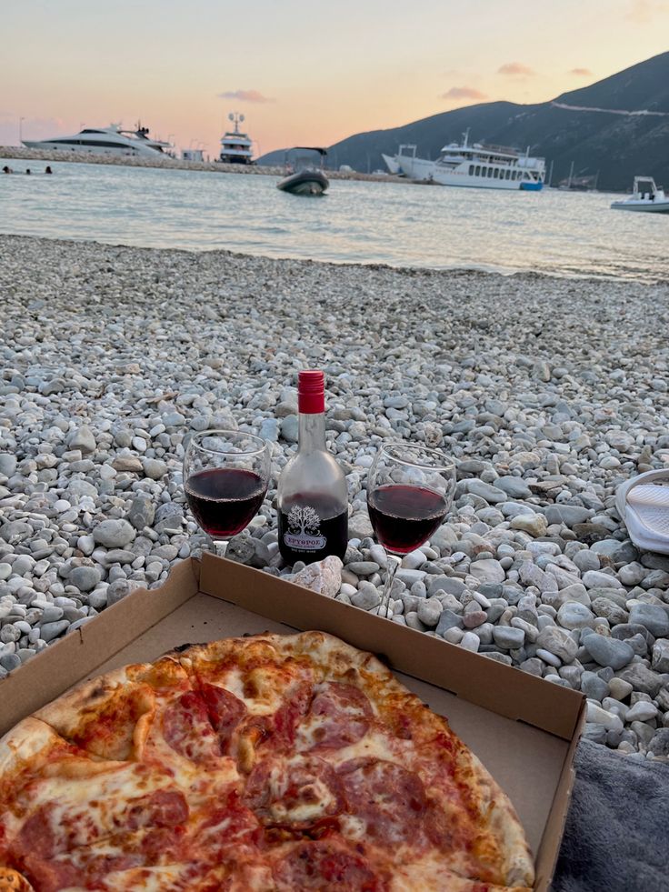 a pizza and two glasses of wine sitting on the beach with boats in the background