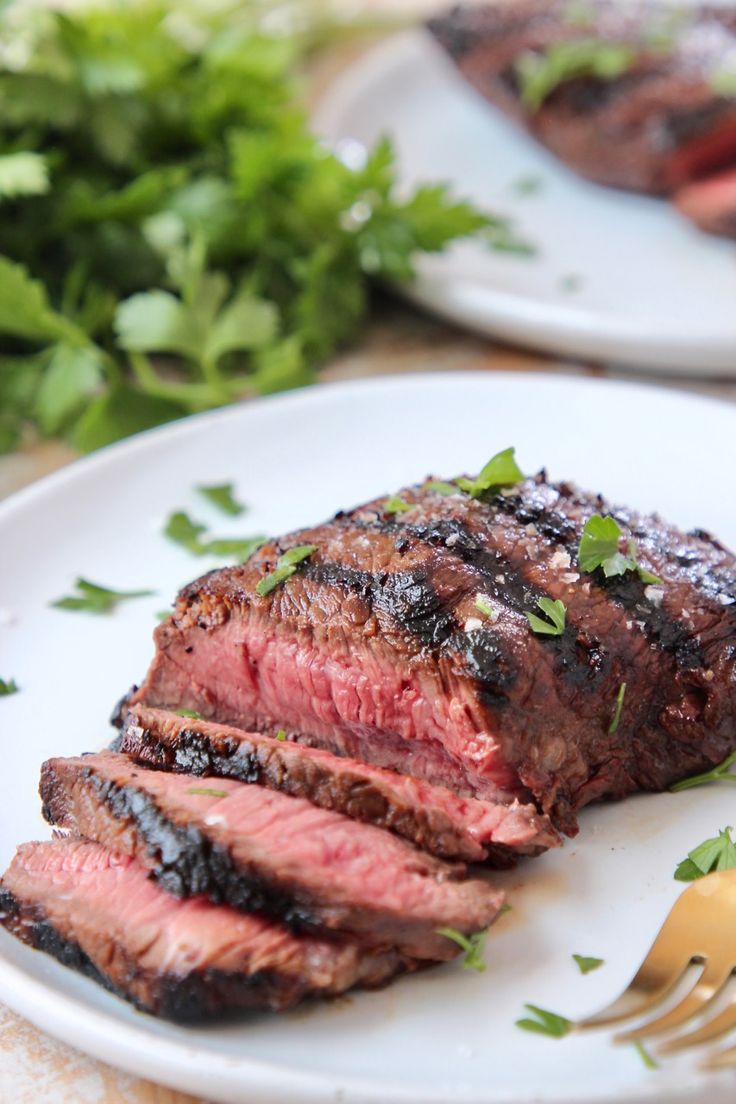 a piece of steak on a white plate with a fork and parsley next to it