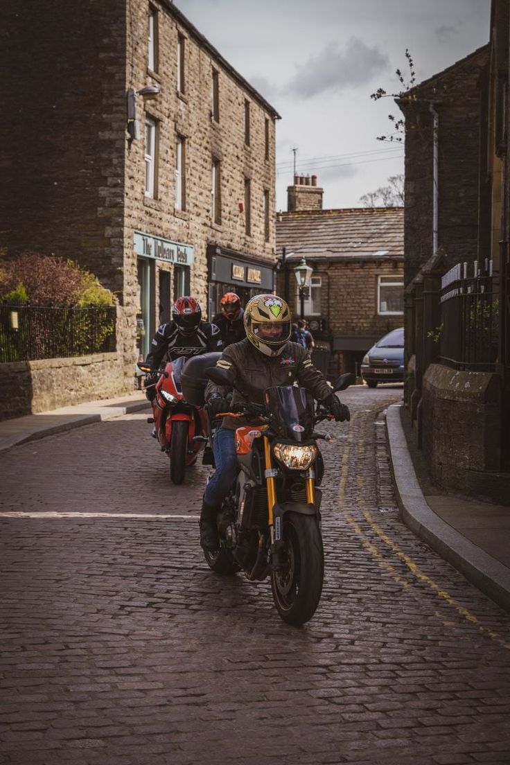 people riding motorcycles down a cobblestone street lined with buildings on either side and one person wearing a helmet