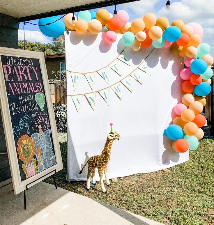 a baby giraffe standing in front of a birthday party sign and balloon arch