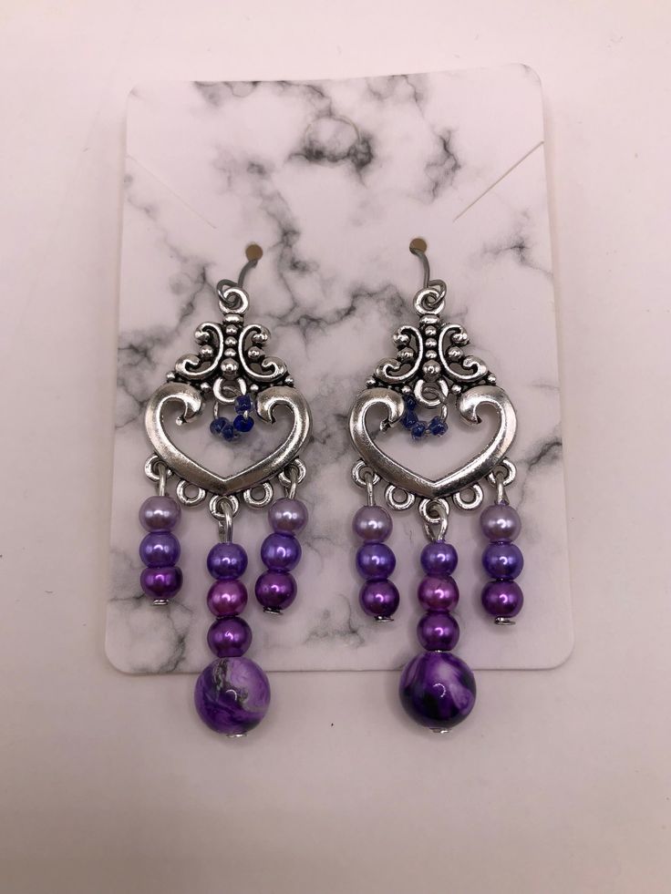 2 Inch drop-earrings made with hypoallergenic materials.   Beads are attached to a silver plated heart charm consisting of a purple marble bead and three smaller glass beads in three different shades of purple/lavender in the center.  Left/Right sides contain the same smaller glass beads in the three different shades of purple/lavender as the center.  The middle of the heart charm is accented with four tiny glass beads as an accent.  These beads are a darker shade of purple. Includes soft ear backings. Materials are environmental, lead free and non-toxic. Light weight, smooth surface, and a touch of sheen to add elegance. The earrings are attached to a marble cardboard earring holder and are shipped in a sealed plastic cover, and placed inside a mesh drawstring "thank you" pouch as shown. Purple Metal Dangle Earrings, Purple Dangle Metal Earrings, Purple Dangle Heart Earrings For Pierced Ears, Purple Metal Earrings With Dangling Beads, Purple Drop Earrings With Dangling Beads, Purple Drop Chandelier Earrings As Gift, Purple Metal Chandelier Earrings For Gifts, Purple Metal Chandelier Earrings As Gift, Purple Dangling Beads Chandelier Earrings